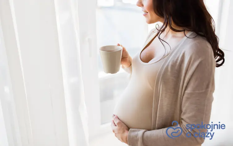 Kobieta w zaawansowanej ciąży z kubkiem przy oknie, a także czy można pić miętę w ciąży