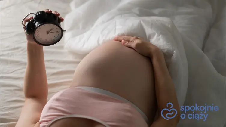 Kobieta w zaawansowanej ciąży leżąca na łóżku i trzymająca budzik, a także czy brak snu w ciąży szkodzi dziecku