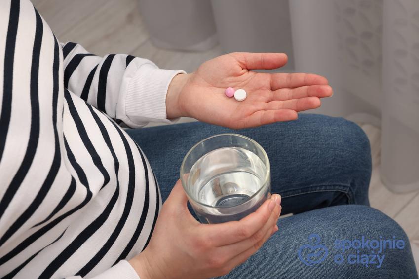Kobieta w ciąży trzymająca pigułki w dłoni oraz szklanę z wodą, a także leki przeciwbólowe