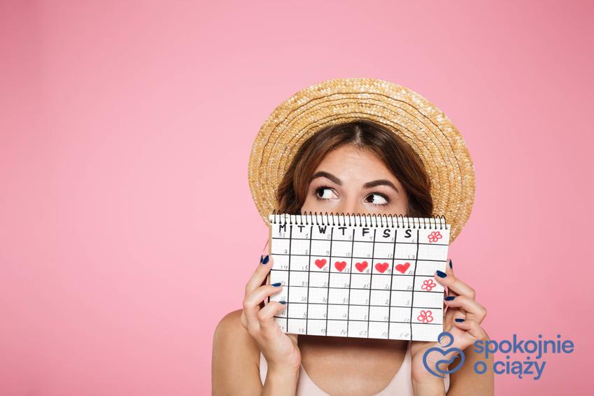 Kobieta w kapeluszu na tle różowej ściany trzymająca kalendarzyk menstruacyjny, a także czy podczas okresu można zajść w ciążę