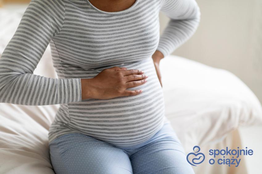 Kobieta w zaawansowanej ciąży siedząca na łóżku i trzymająca się za brzuch, a także ból żołądka w ciąży krok po kroku