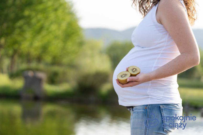 Kobieta w zaawansowanej ciąży trzymająca przekrojone kiwi na wysokości brzucha, a także kiwi w ciąży bez tajemnic