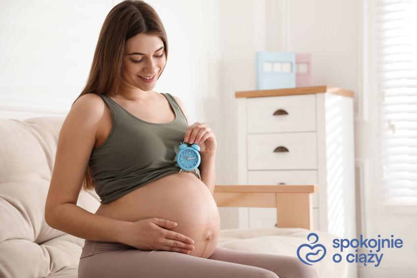 Kobieta w zaawansowanej ciąży trzynająca budzik na wysokości brzucha, a także objawy porodu bez tajemnic