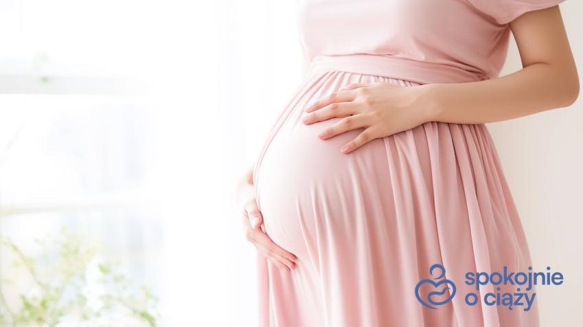 Kobieta w zaawansowanej ciąży w różowej sukience trzymająca się za brzuch, a także informacje, jak zmienia się macica w ciąży